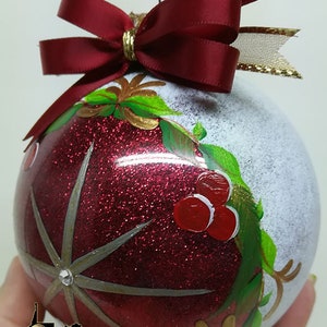 Pode incluir: Um enfeite de Natal vermelho e branco com acabamento de glitter dourado. O enfeite apresenta uma estrela vermelha com contorno prateado e um ramo de azevinho verde com bagas vermelhas. O enfeite é coroado com um laço vermelho e uma fita dourada.