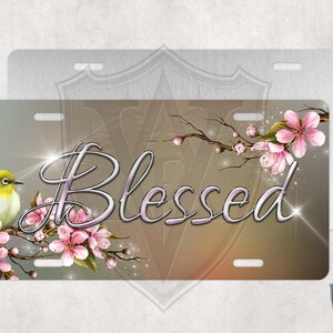 May include: A metal license plate with a light brown background featuring a small yellow bird perched on a branch with pink blossoms. The word "Blessed" is written in a decorative script font.