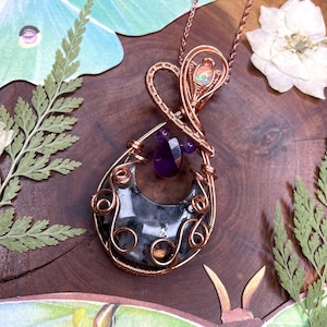 May include: A handcrafted copper wire pendant necklace. The pendant features a teardrop-shaped stone with a dark gray and black pattern, accented with purple amethyst beads and a small opal. The necklace is on a copper chain.