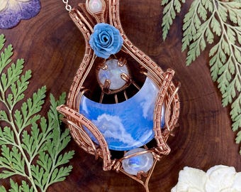 Könnte beinhalten: Ein drahtgewickelter Kupferanhänger mit einem halbmondförmigen blauen Achat, zwei runden Mondsteinen und einer kleinen blauen Rosenverzierung. Das komplexe Kupferdrahtdesign bildet einen tropfenförmigen Rahmen an einer passenden Kupferkette.