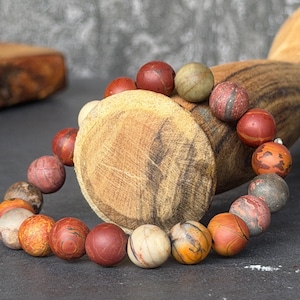 May include: A beaded bracelet featuring spherical stones in various shades of red, orange, tan, and grey. The beads display natural marbling, veining, and earthy textures, arranged in a circular pattern to form a continuous loop of polished mineral spheres.
