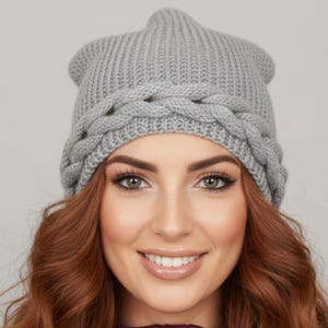 Op de afbeelding: Een grijze gebreide muts met een gevlochten ontwerp rond het voorhoofd. De hoed is gemaakt van een zacht, getextureerd materiaal. De persoon op de foto draagt een bordeauxrode jas met ritssluiting.