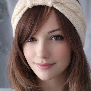 May include: A cream-colored knitted headband with a knotted detail at the top. The headband is worn on a person with brown hair and brown eyes. The knitted texture is visible.