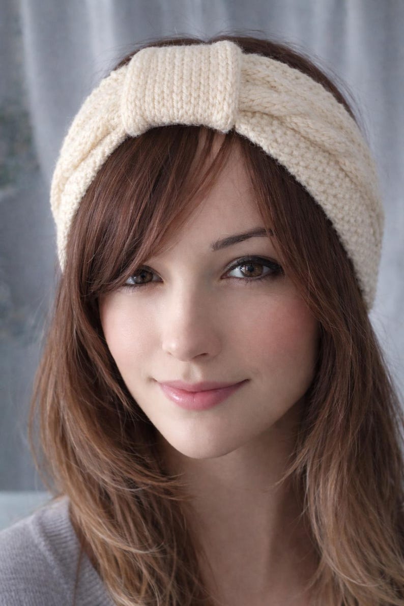 May include: A cream-colored knitted headband with a knotted detail at the top. The headband is worn on a person with brown hair and brown eyes. The knitted texture is visible.