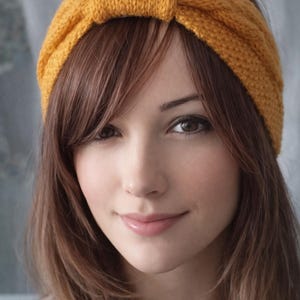 May include: Mustard yellow knitted headband. The headband is wide and features a knotted detail at the top. The accessory is worn on the head and frames the face. The hair is long and brown.
