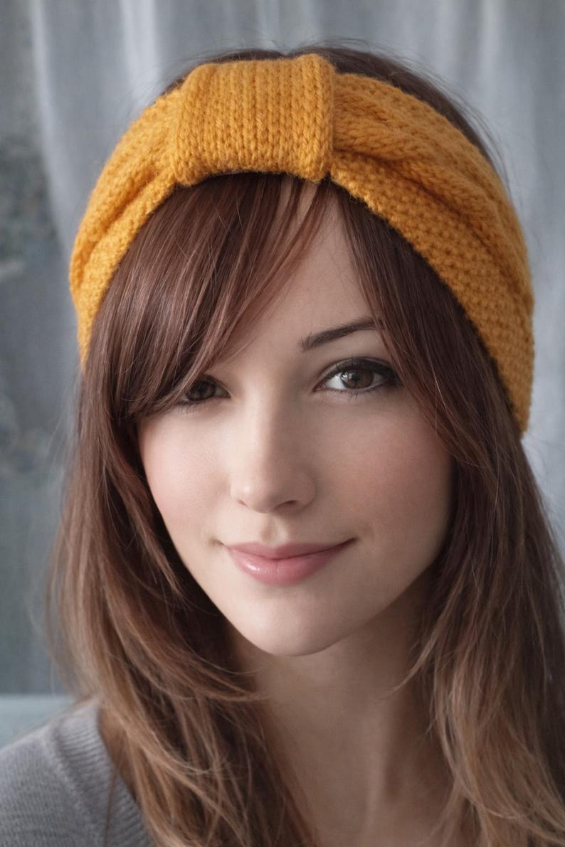 May include: Mustard yellow knitted headband. The headband is wide and features a knotted detail at the top. The accessory is worn on the head and frames the face. The hair is long and brown.