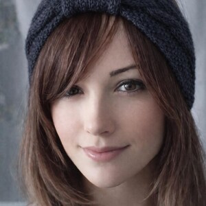 May include: A dark blue knitted headband with a knotted detail. The headband is worn on the head, with brown hair framing the face. The person is wearing a gray top.