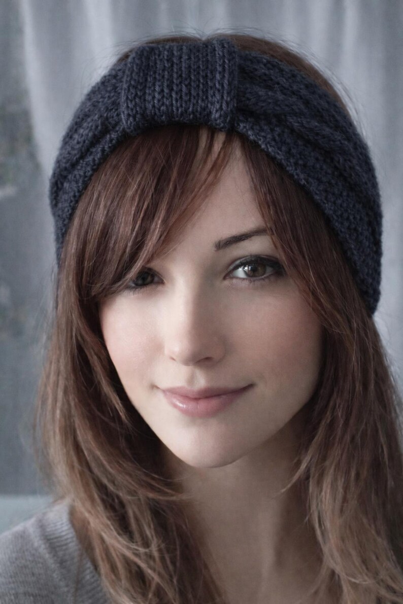 May include: A dark blue knitted headband with a knotted detail. The headband is worn on the head, with brown hair framing the face. The person is wearing a gray top.