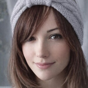 May include: A light gray knitted headband with a knotted detail. The headband is worn on the head, with brown hair framing the face. The image is a close-up, showing the texture of the knit and the person's features.