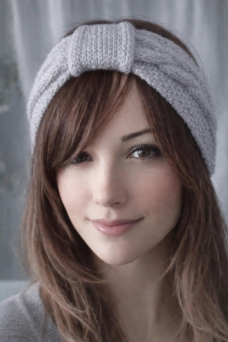 May include: A light gray knitted headband with a knotted detail. The headband is worn on the head, with brown hair framing the face. The image is a close-up, showing the texture of the knit and the person's features.