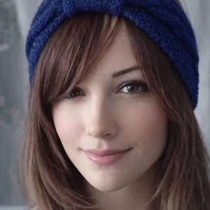 May include: A close-up of a woman wearing a knitted blue headband. The headband is a solid, vibrant blue color and has a textured appearance. The woman has long, light brown hair and is looking at the camera.