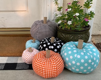 Puede incluir: Seis calabazas de tela en varios patrones y colores. Las calabazas están dispuestas en una alfombra de bienvenida frente a una puerta blanca. Las calabazas son de cuadros blancos y negros, de lunares blancos y negros, de color azul claro con lunares blancos, de cuadros blancos y naranjas, y dos calabazas más pequeñas de cuadros blancos y naranjas.