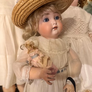 May include: A vintage porcelain doll with blonde hair and blue eyes, wearing a straw hat and a white dress with lace trim. The doll holds a smaller, miniature doll dressed in a pink and white outfit. The doll has a pale complexion and rosy cheeks.
