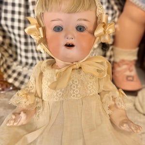 May include: A porcelain doll with blonde hair, blue eyes, and an open mouth. The doll wears a cream-colored dress with lace trim and a matching bonnet tied with a ribbon under the chin. The doll has pale skin and articulated hands.