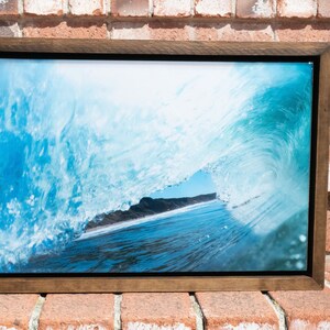 Puede incluir: Una fotografía enmarcada de una gran ola rompiendo, mostrando el océano a través de la cresta de la ola. La ola es de un azul y blanco vibrantes, con un vistazo del cielo azul y una playa de arena en la distancia.