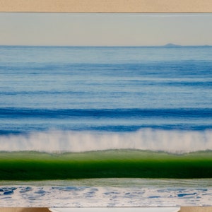 May include: A photograph of a large wave crashing on a beach. The wave is green and white, and the ocean behind it is blue. The horizon is in the distance, and there is a small island in the distance.