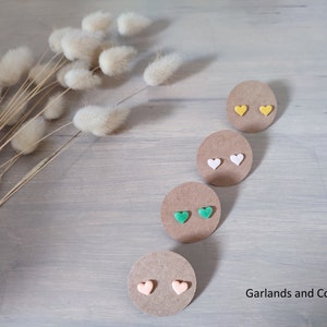 Peut inclure: Quatre paires de boucles d'oreilles en forme de cœur présentées sur des cartes rondes en carton brun. Les boucles d'oreilles sont de couleur jaune, blanche, verte et pêche. Le texte Garlands and Co est imprimé dans le coin inférieur droit.