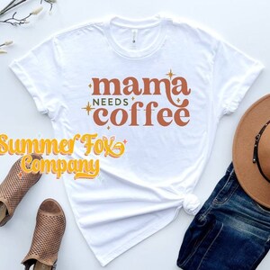 Könnte beinhalten: Weißes T-Shirt mit dem Text "mama needs coffee" in orangefarbener und goldener Schrift mit Sternen. Das Shirt ist mit einem braunen Hut, einer blauen Jeans und braunen Sandalen gestylt.