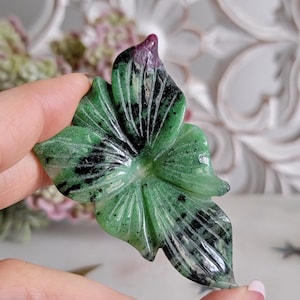 May include: A green and black carved flower pendant made from green aventurine gemstone. The flower has a pink center and is carved with intricate details.