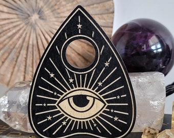 Puede incluir: Tablero de Ouija de madera negra con un ojo blanco y un diseño de estrellas. El tablero tiene forma de lágrima y tiene un agujero en el centro.