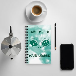 May include: A spiral-bound notebook with a teal tie-dye design and the text "TAKE ME TO YOUR LEADER." The notebook features an alien face graphic. A cup of coffee, a pencil, a phone, and a stovetop espresso maker are also on the white surface.