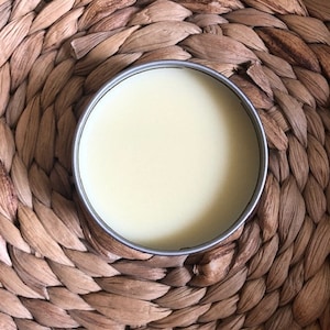 May include: A round, silver-rimmed container filled with a pale yellow substance, possibly a balm or cream. The container sits on a woven, brown, textured surface, creating a natural and earthy aesthetic. The product is presented in a simple, minimalist style.