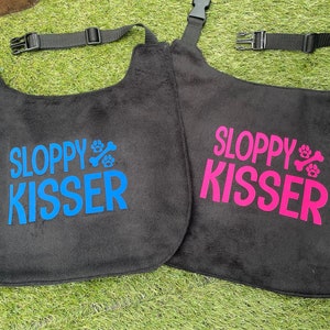 May include: Two black dog bibs with adjustable straps. One bib has the text "Sloppy Kisser" in blue with paw prints and a bone. The other bib has the text "Sloppy Kisser" in pink with paw prints and a bone.