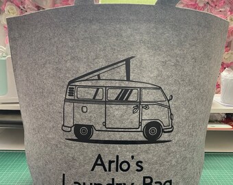 May include: A gray felt laundry bag with two handles. The bag features a black graphic of a camper van and the text "Arlo's Laundry Bag" in black. The bag is cylindrical and sits on a green surface.