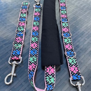 Könnte beinhalten: Hundeleinen-Set mit einem schwarzen, rosa, blauen und grünen Pfotenabdruckmuster auf weißem Hintergrund mit weißen Punkten. Das Set enthält eine Leine mit gepolstertem Griff und zwei zusätzliche Leinenbefestigungen mit Metallverschlüssen und Ringen.