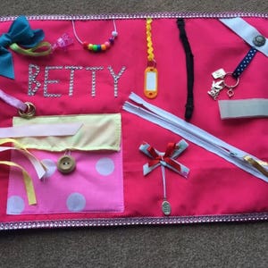 May include: A pink sensory board with various textures and objects for tactile exploration. Features include a teal bow, a beaded necklace, a zipper, a button, and the name "BETTY" spelled out in rhinestones. The board is trimmed with a sparkly silver border.
