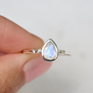 May include: A silver ring featuring a pear-shaped moonstone set in a bezel setting. The ring has a delicate, textured band.