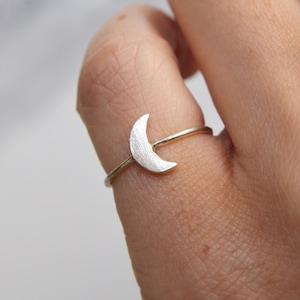 Könnte beinhalten: Ein silberner Ring mit einem Mondmotiv. Der Mond hat eine strukturierte Oberfläche und sitzt auf einem schmalen Band. Der Ring wird an einem Finger gezeigt, mit einem unscharfen Hintergrund.