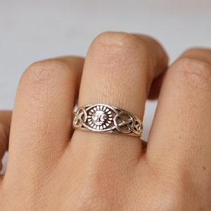 Puede incluir: Un anillo plateado con un diseño de sol en el centro, rodeado de intrincados patrones de nudos celtas. El anillo se lleva en un dedo, mostrando su detallada artesanía y su acabado metálico brillante. El diseño del anillo es simétrico y detallado.