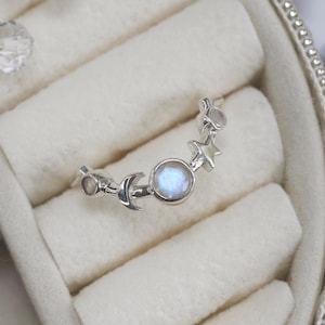 以下が含まれることがあります： 月と星のデザインが施されたシルバーリング。中央にはムーンストーン、三日月、星が配置されています。白いベルベットのジュエリーボックスに収められ、背景にはパールのアクセントがあります。