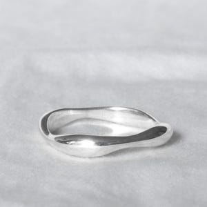May include: A silver ring with a flowing, organic design. The ring has a smooth, polished surface and a gently undulating shape. The ring is set against a soft, white background, highlighting its minimalist aesthetic.