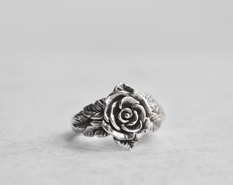 May include: A silver ring with a detailed rose design. The rose is centered on the ring and has leaves extending out from the sides.