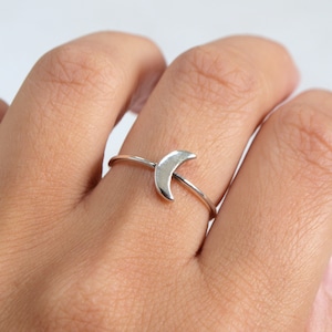 May include: A silver ring with a crescent moon design. The ring has a thin band and a small moon shape. The ring is worn on a finger, highlighting its simple and elegant style. The ring is a piece of jewellery, suitable for everyday wear.