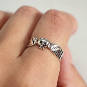 May include: A silver ring featuring a skull with feathered wings. The ring is detailed with a skull face and wing design, and is worn on a finger. The ring is a gothic or fantasy style.