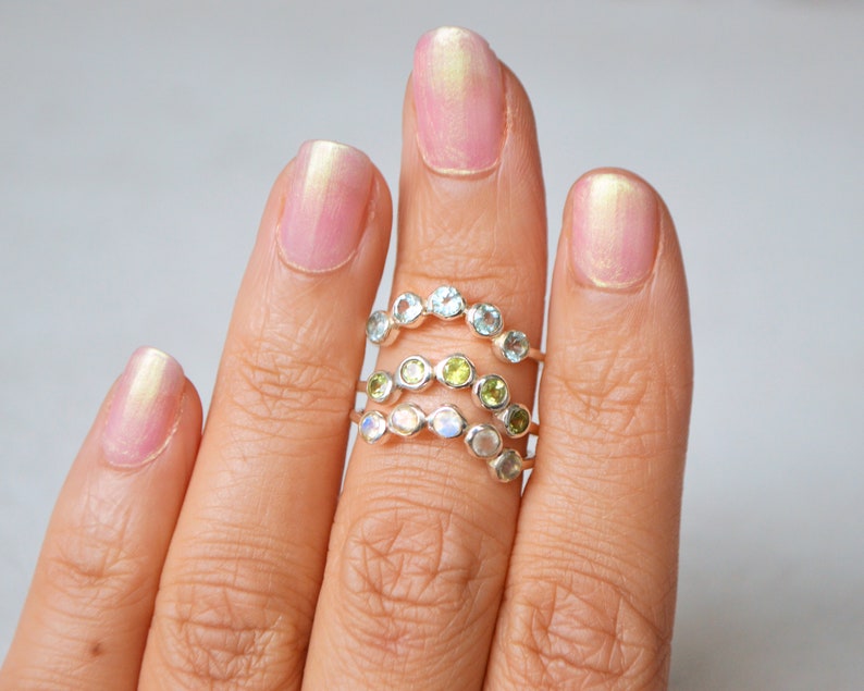 May include: Three silver rings with different coloured gemstones. The top ring has light blue gemstones, the middle ring has green gemstones, and the bottom ring has white gemstones.