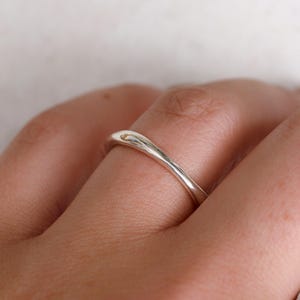 Könnte beinhalten: Ein Ring aus poliertem Silber mit einer glatten, wellenförmigen Ringschiene. Das Metall hat eine reflektierende, hochglänzende Oberfläche, die das Licht einfängt und subtile Glanzlichter entlang der geschwungenen Oberfläche erzeugt.