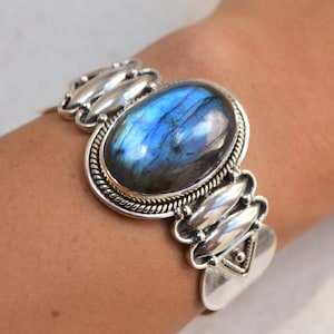 Puede incluir: Pulsera de brazalete de plata con una gran piedra ovalada de labradorita azul. La pulsera tiene un diseño detallado con detalles plateados.