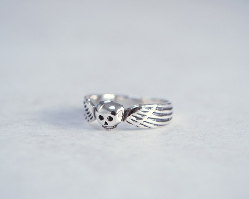 May include: A silver ring featuring a skull with wings. The skull has two small eye sockets and a detailed jawline. The wings extend from the sides of the skull, with detailed feathering. The ring is a unique piece of jewelry.