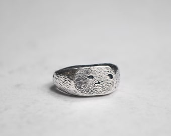 May include: A silver ring with a textured, oval-shaped face featuring a stylized, abstract design.