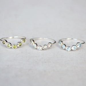 May include: Three silver rings each featuring a row of gemstones. The rings are arranged in a row from left to right. The ring on the left has green gemstones, the middle ring has light blue gemstones, and the ring on the right has dark blue gemstones.