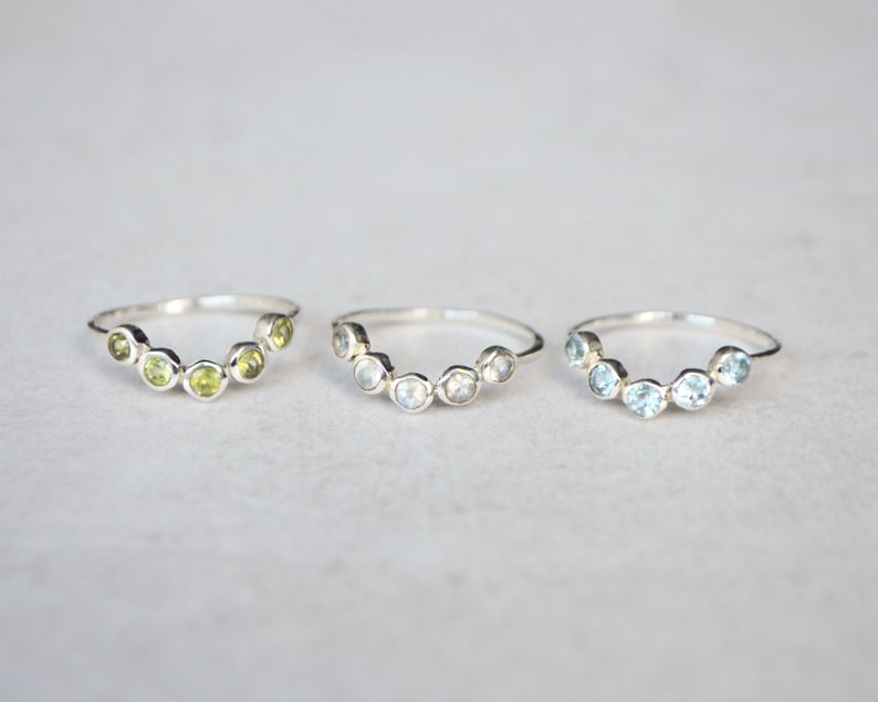 May include: Three silver rings each featuring a row of gemstones. The rings are arranged in a row from left to right. The ring on the left has green gemstones, the middle ring has light blue gemstones, and the ring on the right has dark blue gemstones.