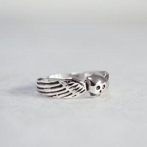 May include: A silver ring featuring a skull with wing accents. The ring has a detailed design, with the skull's features clearly visible. The wings are on either side of the skull. The ring is likely made of sterling silver.