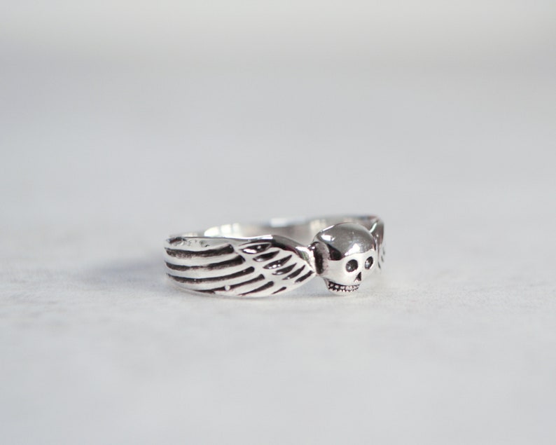 May include: A silver ring featuring a skull with wing accents. The ring has a detailed design, with the skull's features clearly visible. The wings are on either side of the skull. The ring is likely made of sterling silver.