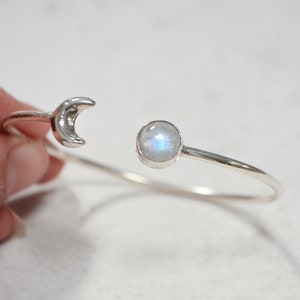 Puede incluir: Una pulsera abierta de plata con un dije de media luna en un extremo y un cabujón de piedra lunar redondo e iridiscente engastado en un bisel de plata en el otro. La banda de metal es delgada, pulida y tiene un diseño circular.