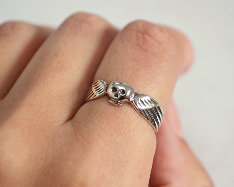 May include: A silver ring featuring a skull with feathered wings. The ring is detailed with a skull face and wing design, and is worn on a finger. The ring is a gothic or fantasy style.
