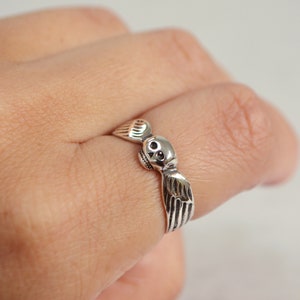 May include: A silver ring featuring a skull with detailed wings. The ring is worn on a finger, showcasing the intricate design and craftsmanship. The skull has detailed eye sockets and a small nose.